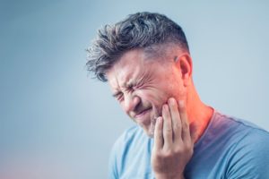 man with gray hair in blue shirt squinting in pain and pressing fingers to aching jaw